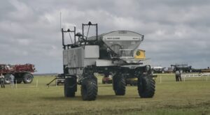Máquina agrícola em campo aberto com pessoas e outras máquinas ao fundo.